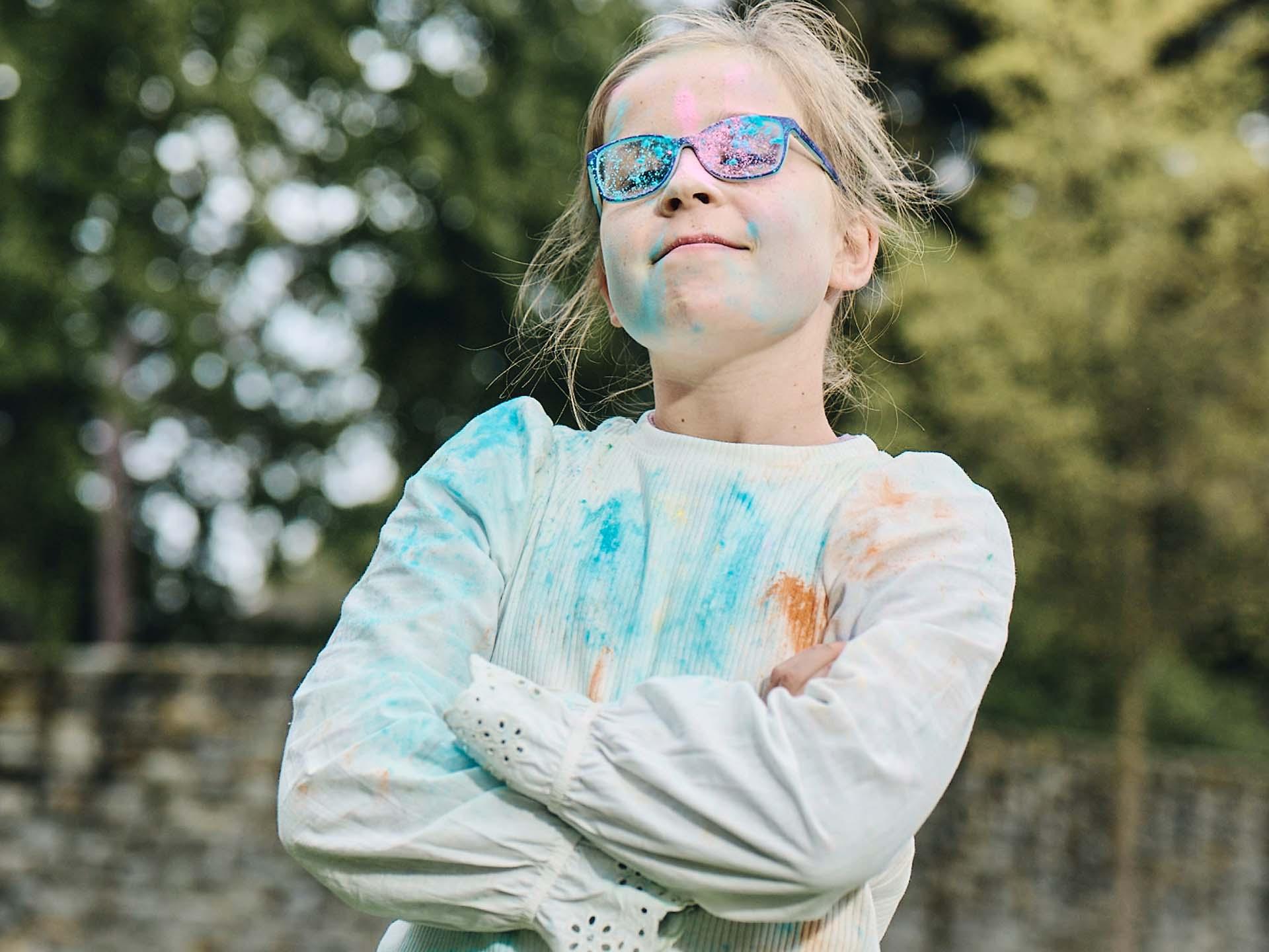 Dopo aver giocato con la polvere colorata, una bambina con le braccia incrociate e gli occhiali sporchi guarda coraggiosa e sorride.