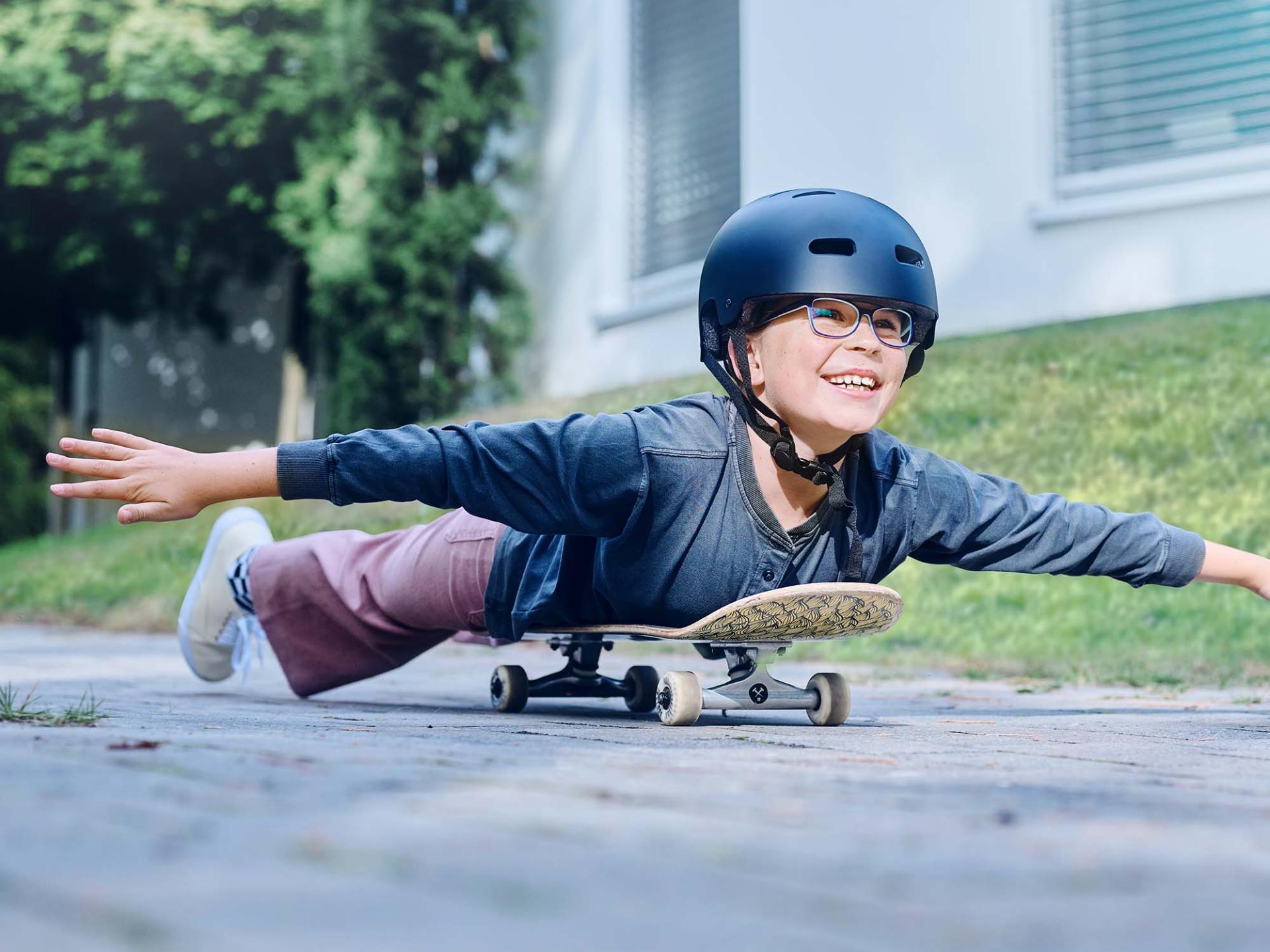 Una bambina con casco e occhiali percorre la strada su uno skateboard e allunga le braccia.