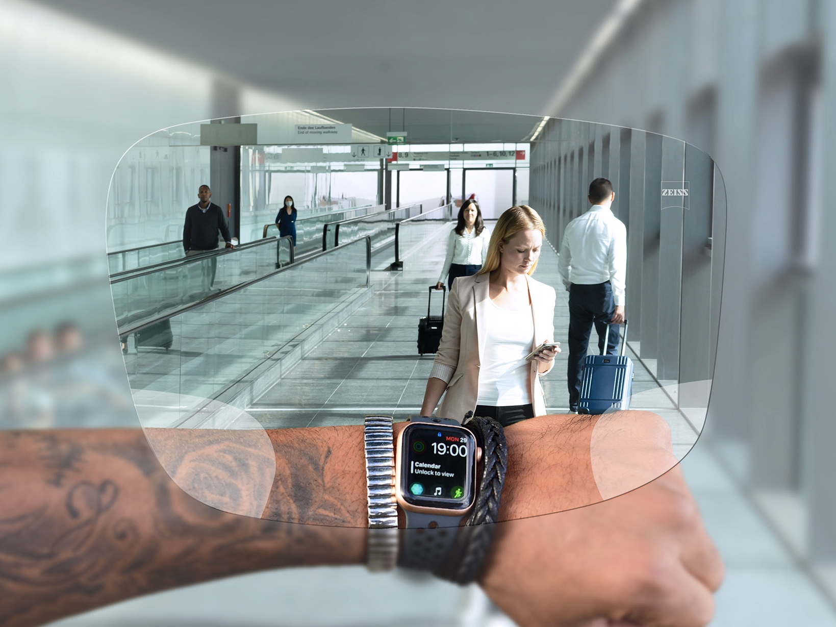 Una persona guarda il suo orologio attraverso le lenti ZEISS SmartLife Digital. Sullo sfondo si vede la situazione tipica di un aeroporto con persone che camminano sulle scale mobili e trascinano le valigie. 